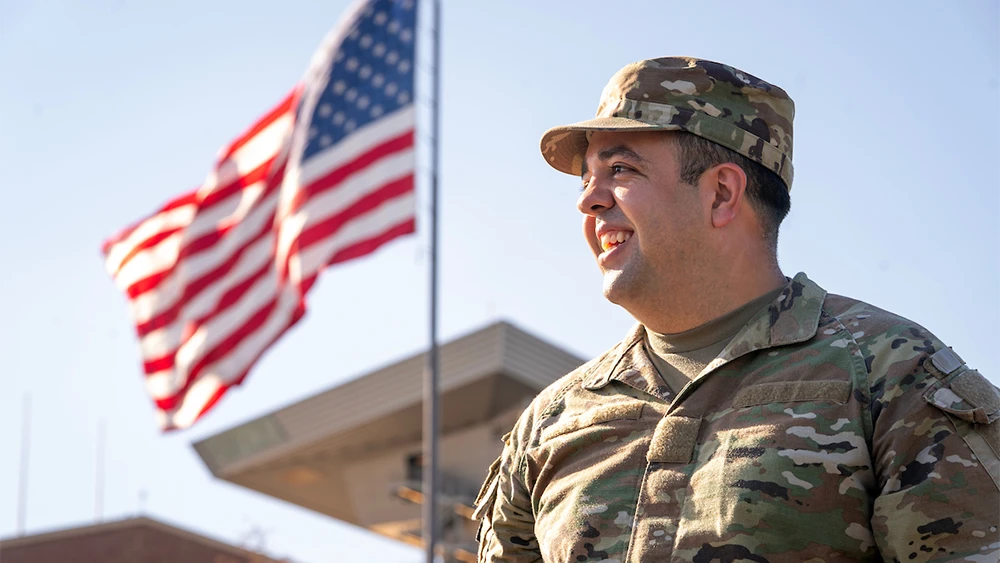 A veteran in military fatigues smiles in front of a waving American flag