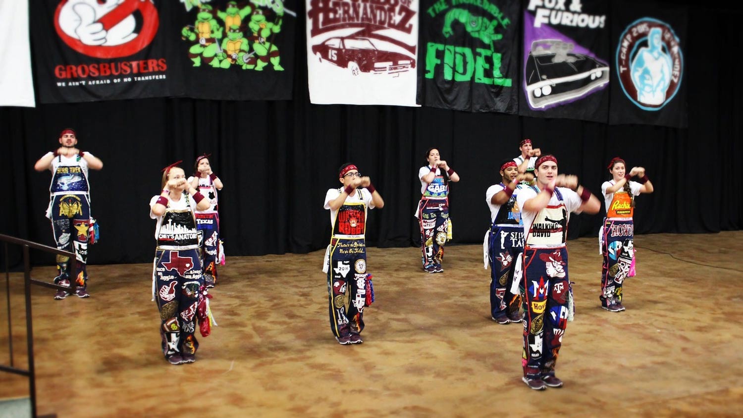 A group of camp leaders in decorated overalls on stage at T-Camp