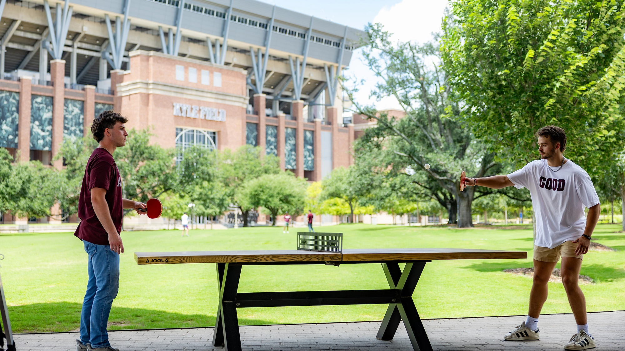Two college students playing table tennis outdoors in front of a large sports stadium.
