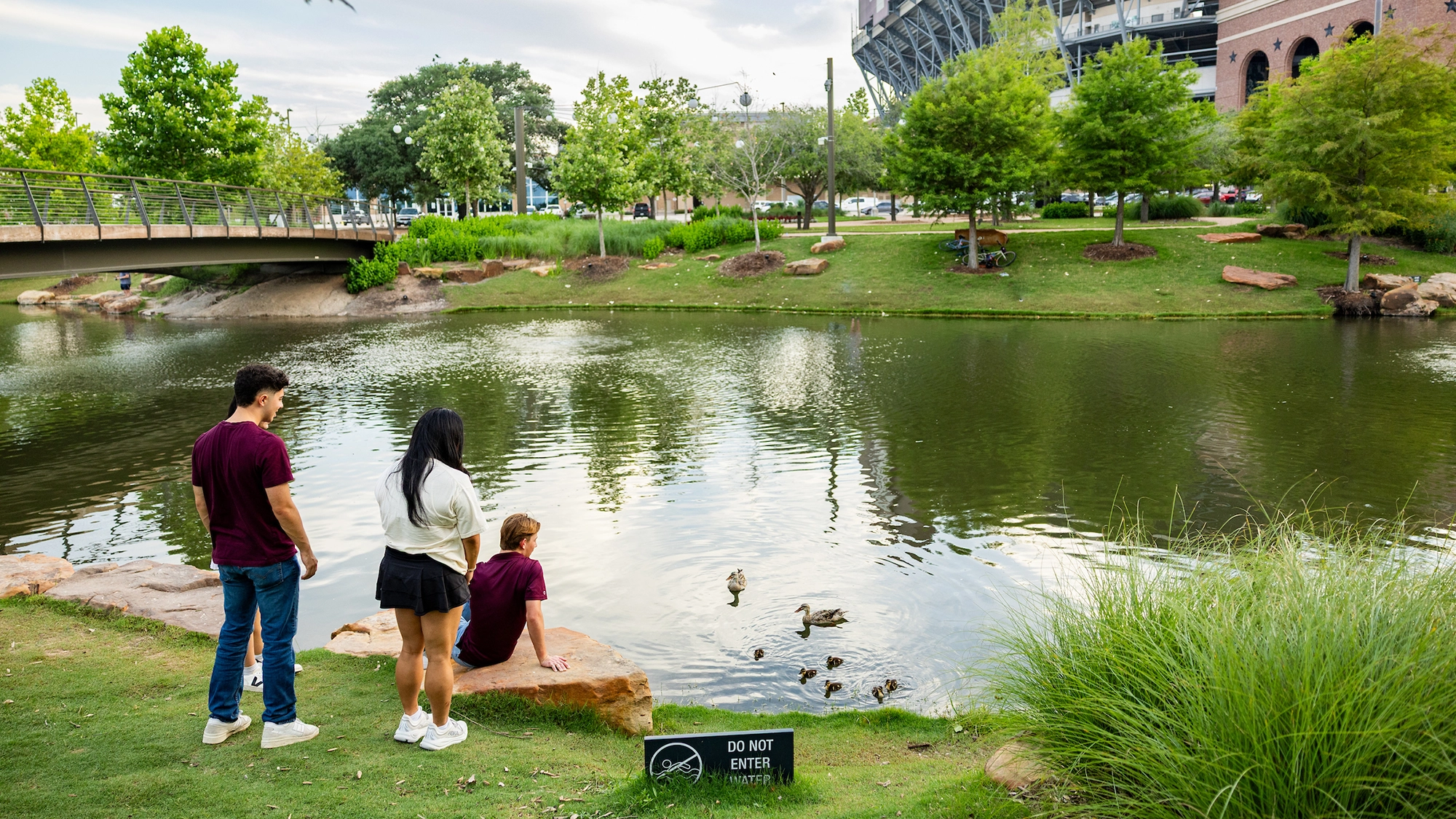 A group of three people, wearing casual outfits, observe ducks on a serene pond surrounded by greenery.