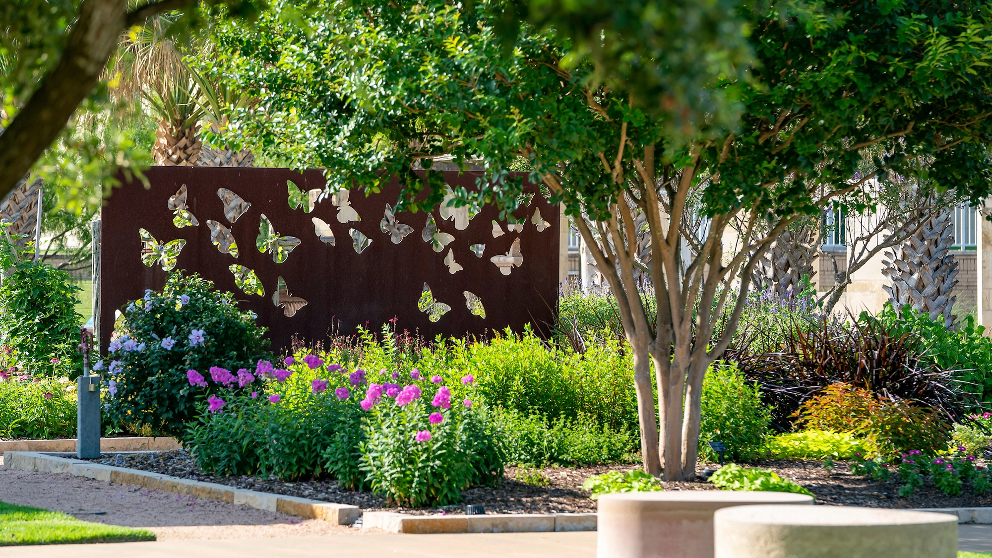 A decorative metal screen featuring butterfly cutouts surrounded by vibrant flowers and greenery in a landscaped garden.