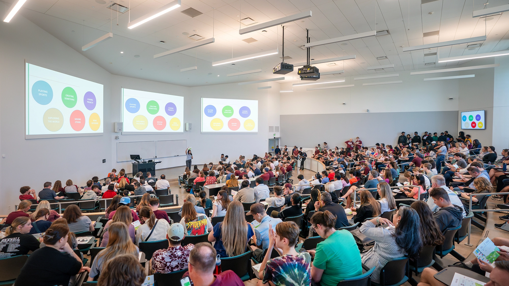 A large lecture hall filled with students attending a presentation, featuring colorful slides displayed on two screens.